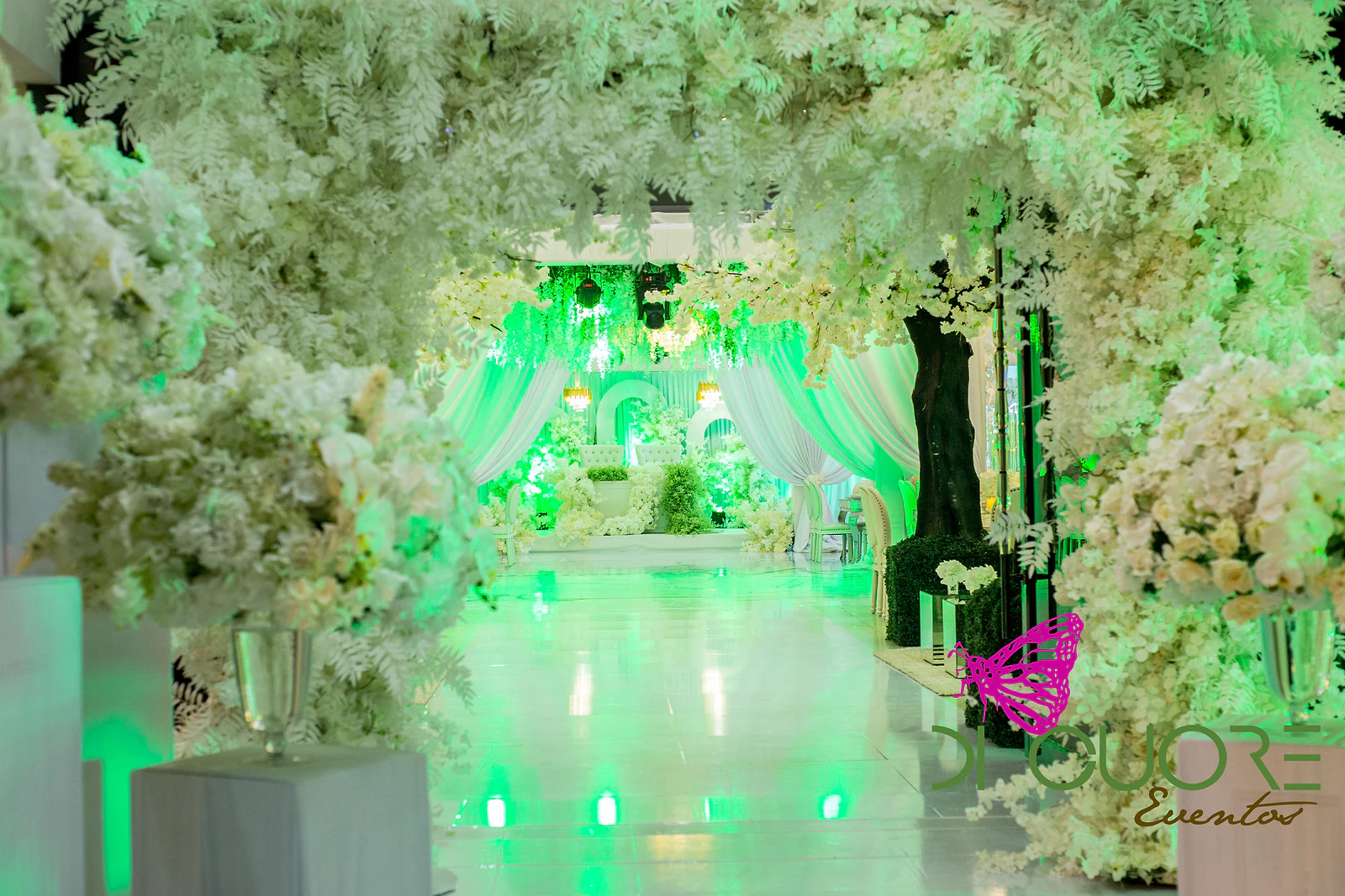 Passagem sob um arco de flores brancas e folhagem, entre pedestais com grandes ramos, a conduzir ao palco ao fundo
