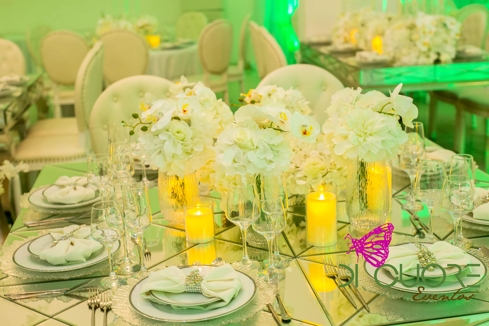 Mesa de espelho com três ramos de hortênsias e orquídeas em vasos de vidro, velas e guardanapos presos por argolas de cristal