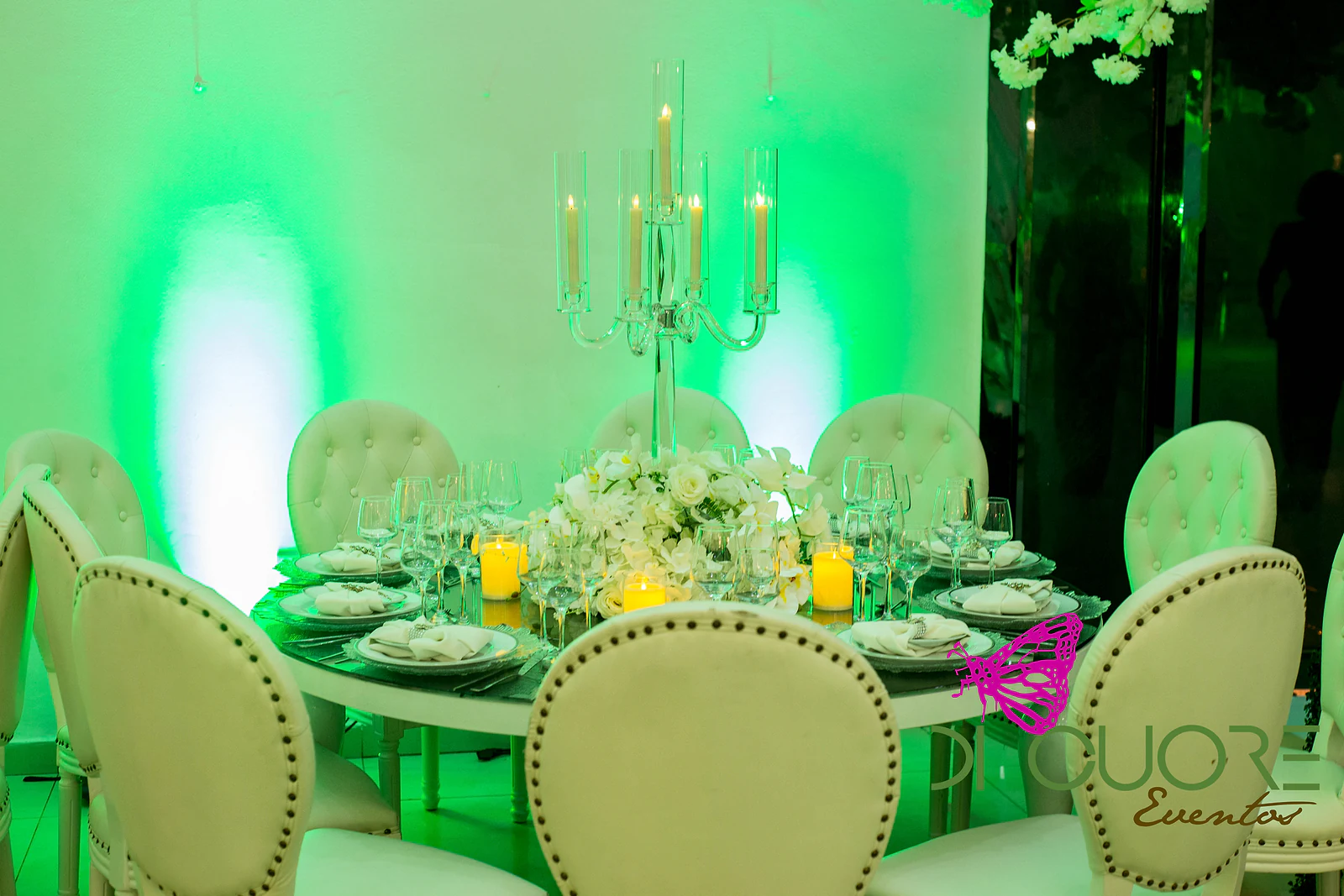 Mesa redonda de espelho com ramo de rosas e orquídeas, velas e candelabro de vidro, rodeada de cadeiras brancas de botões, diante de uma parede iluminada