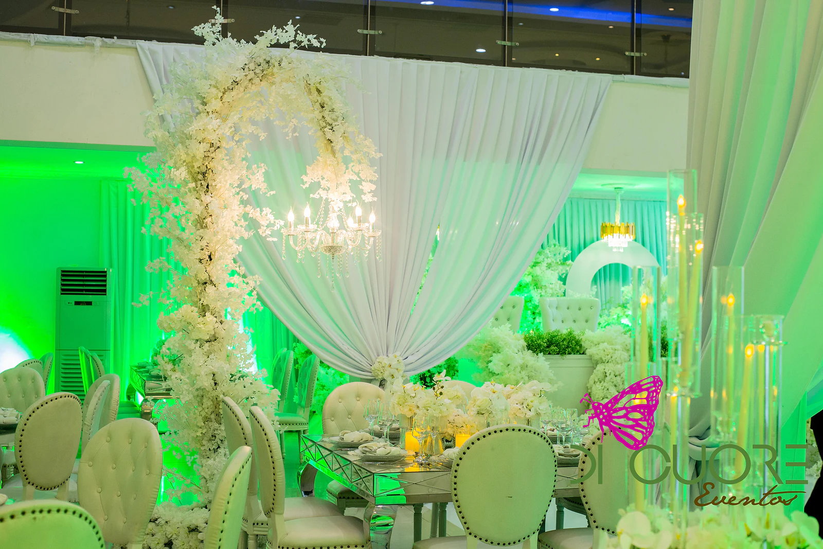 Arco de flores brancas com um candelabro de cristal suspenso, diante de um grande cortinado branco, sobre mesas postas