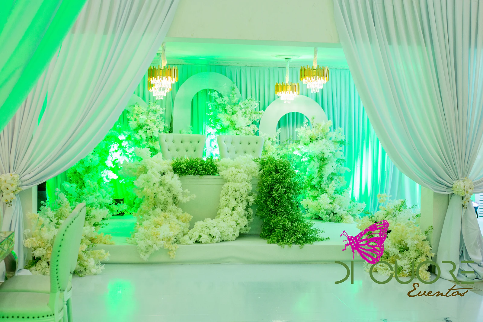 Palco dos noivos entre cortinados brancos atados com flores, com dois cadeirões capitonados, arcos brancos, lustres dourados e cascata de flores e folhagem