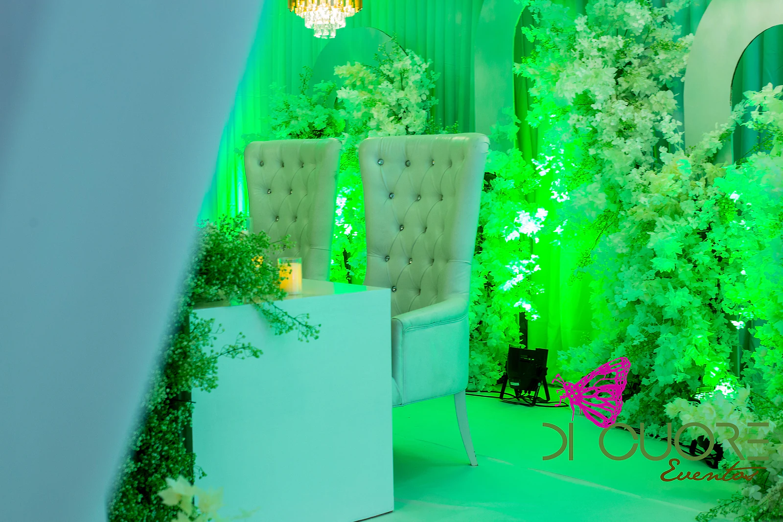 Dois cadeirões brancos de encosto capitonado com botões de cristal, junto a um balcão branco com folhagem e uma vela, entre ramos de flores brancas