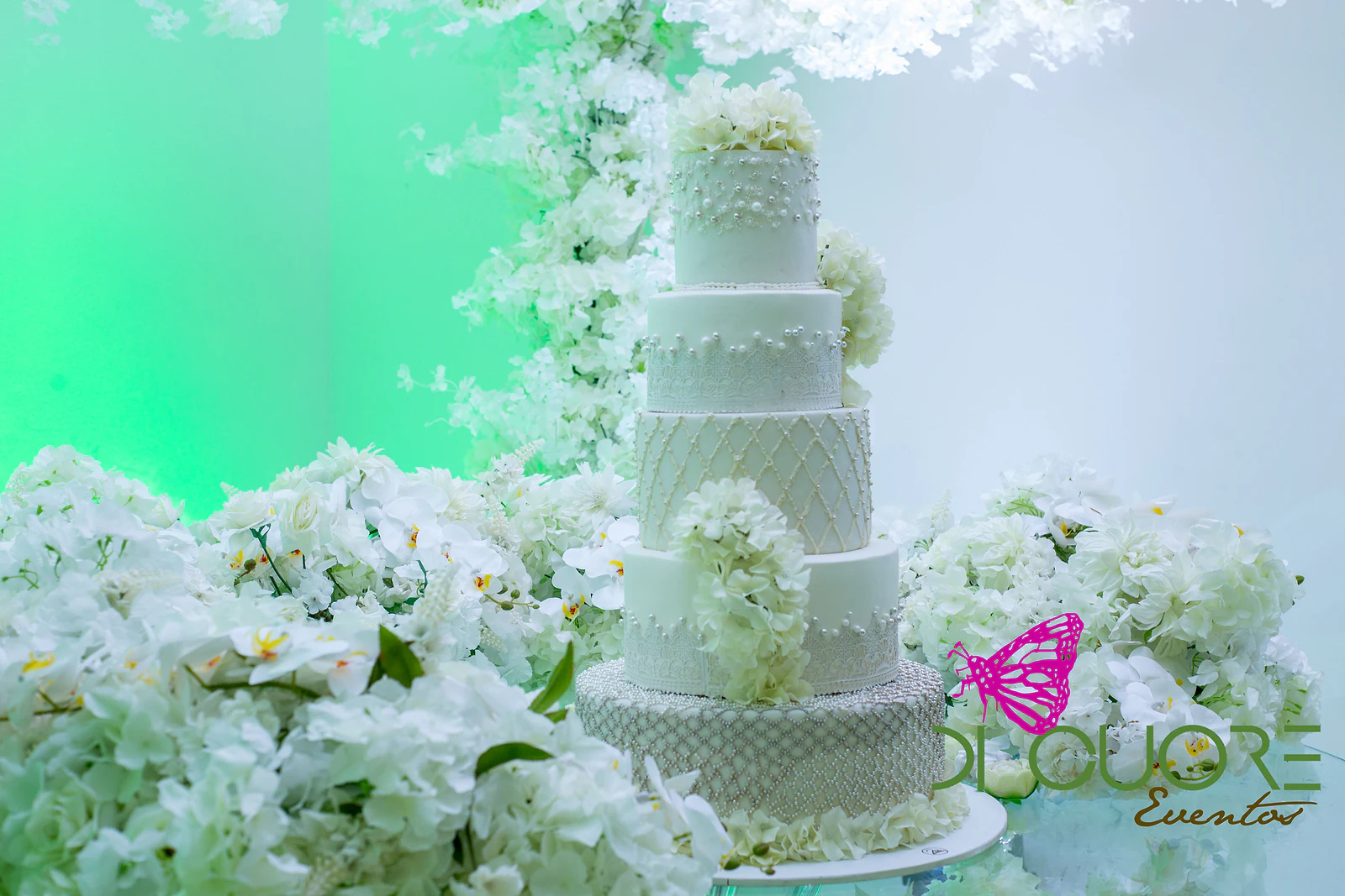 Bolo de casamento de cinco andares com padrão em losango e hortênsias, rodeado de hortênsias e orquídeas brancas