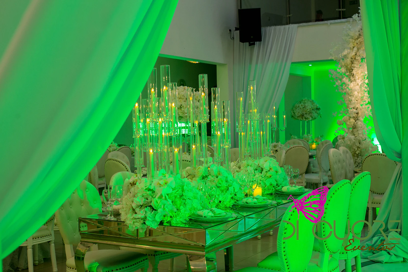 Mesa de espelho com ramos brancos e uma fileira de candelabros altos de vidro com velas, entre cadeiras claras e cortinado verde