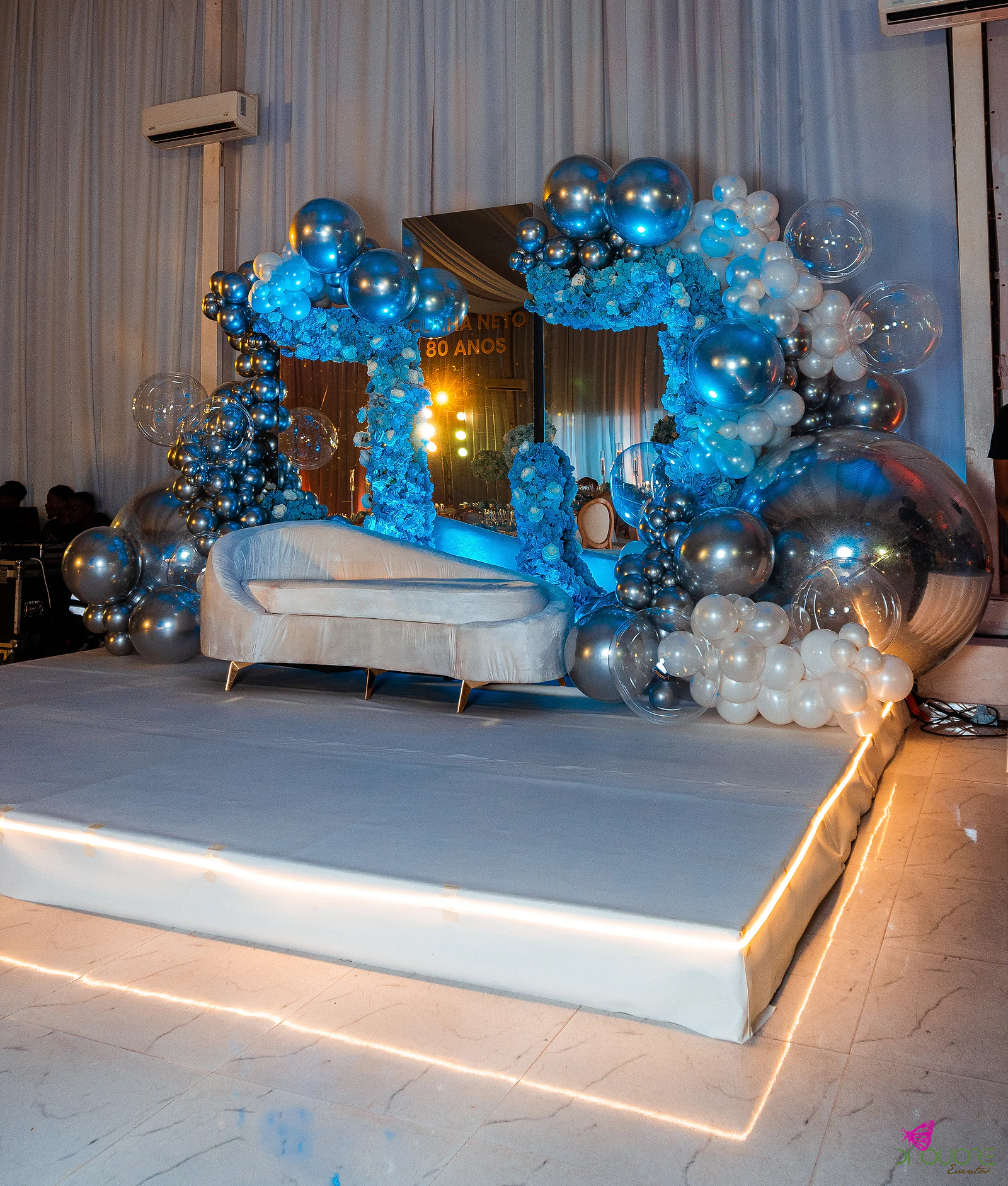 Palco elevado com rebordo luminoso, chaise longue branco e arco de balões prateados com flores azuis