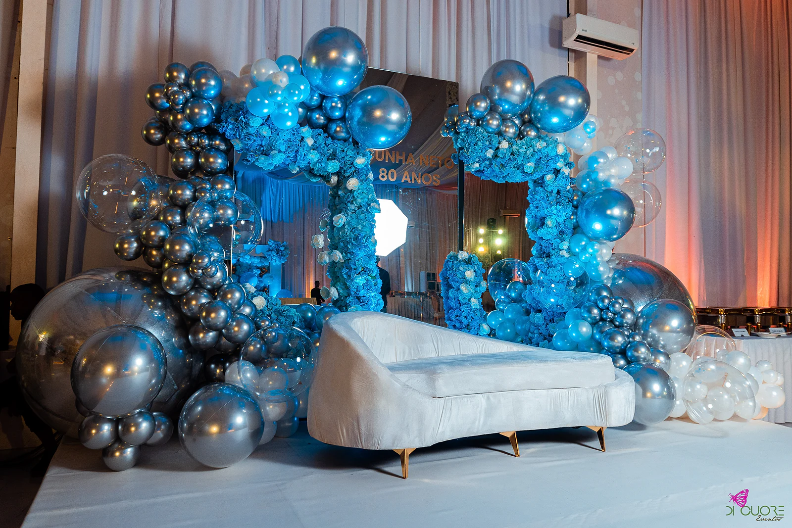 Chaise longue branco diante de arcos de balões prateados e azuis com flores, sobre um palco de cortinados claros