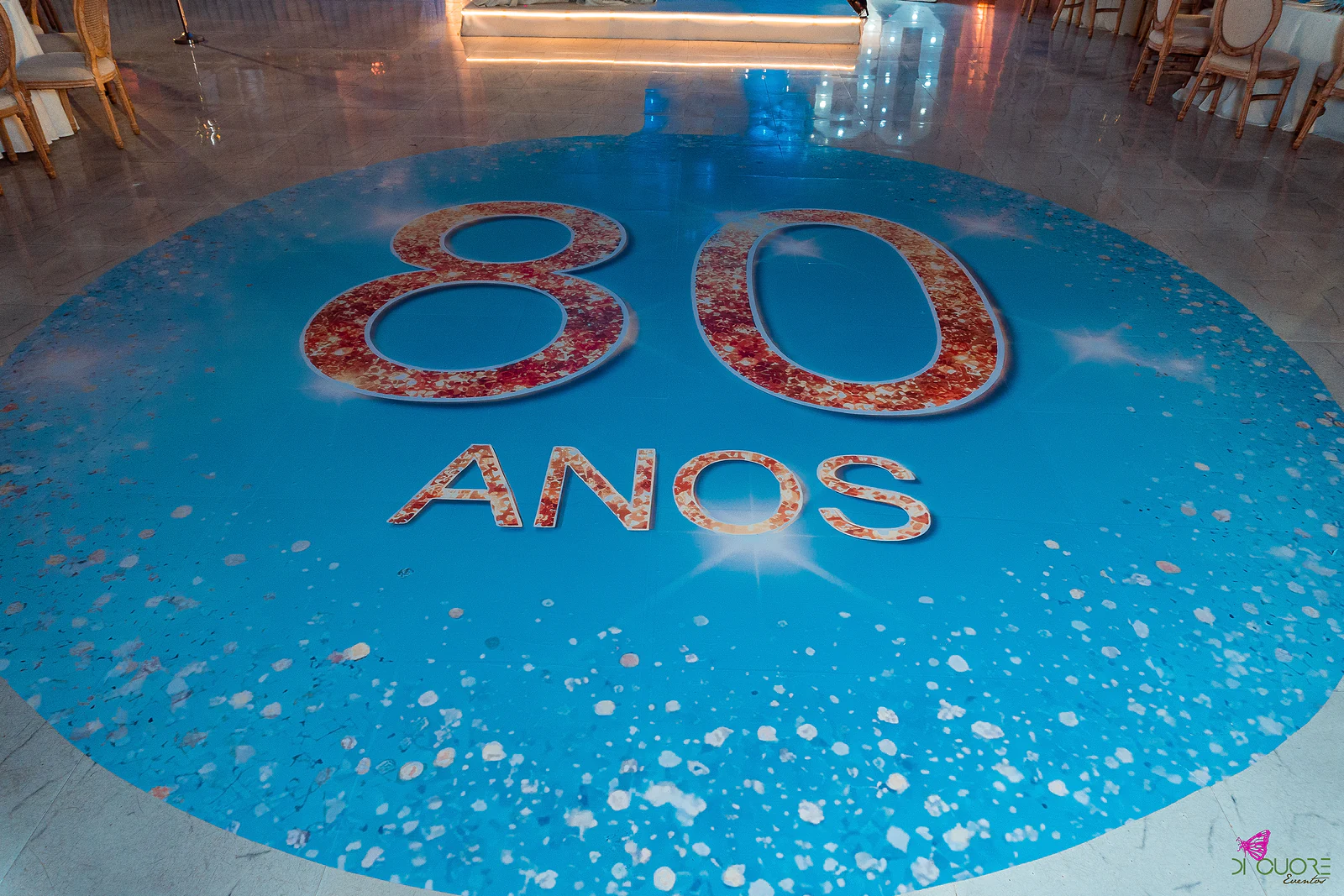 Círculo azul no chão com o número 80 e a palavra ANOS em letras rosadas, salpicado de pétalas brancas, com o estrado iluminado ao fundo