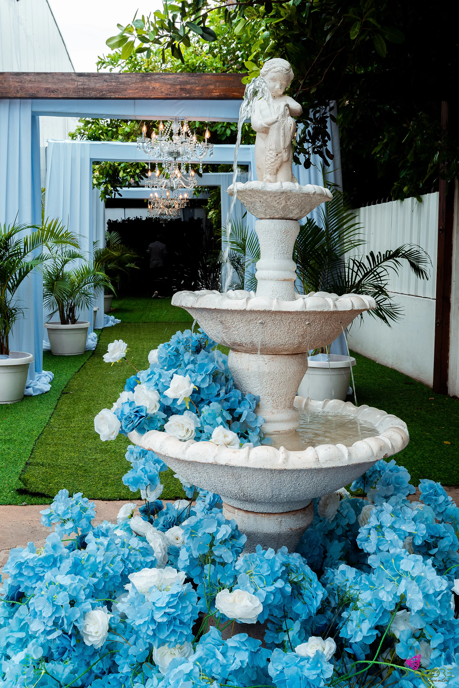 Fonte de três taças com um anjo de pedra, rodeada de hortênsias azuis e rosas brancas, junto à relva