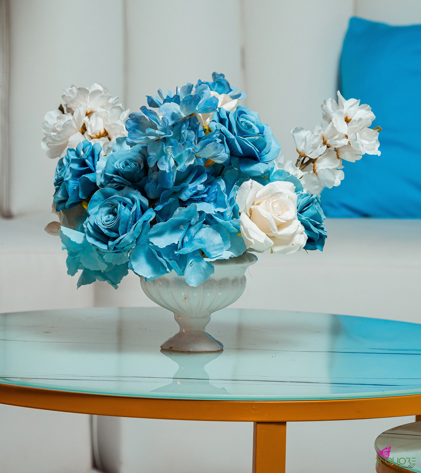 Taça branca com rosas e flores em azul e branco sobre uma mesa baixa de tampo de vidro azul-claro e pé dourado