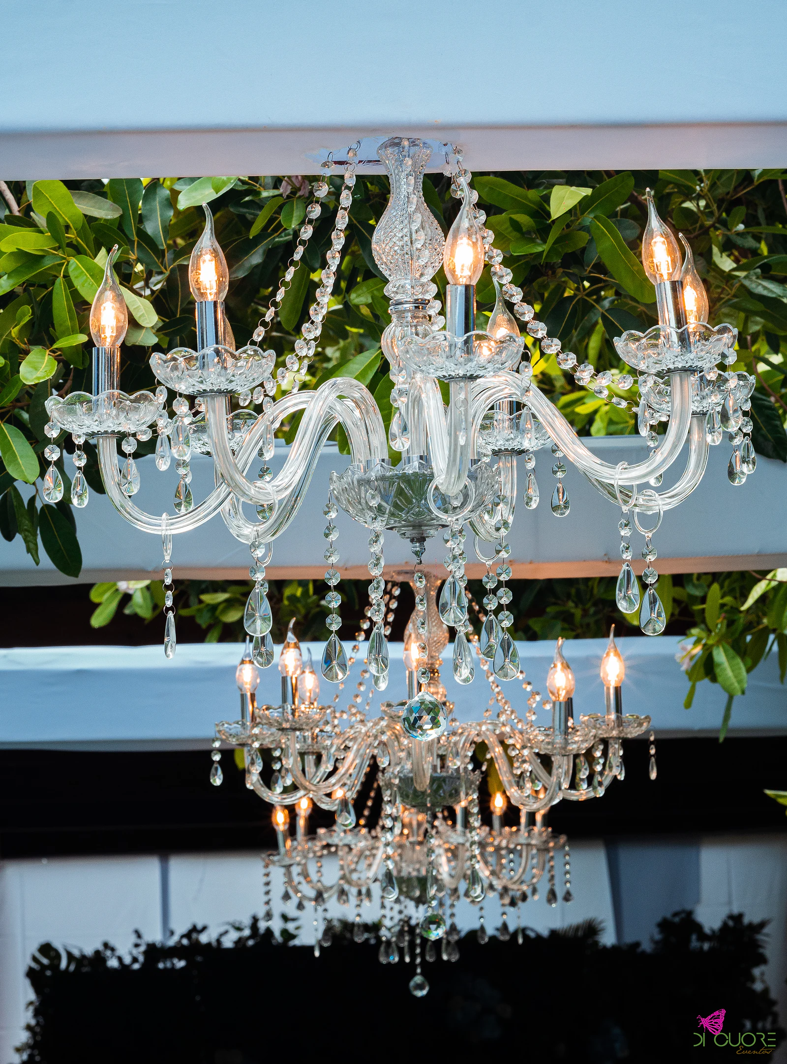 Lustre de cristal com velas pendurado entre folhagem, num pórtico de cortinados brancos