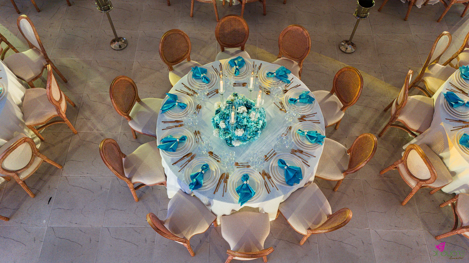 Mesa redonda vista de cima, com guardanapos azuis nos pratos, velas e um arranjo de hortênsias azuis e brancas ao centro, rodeada de cadeiras de madeira