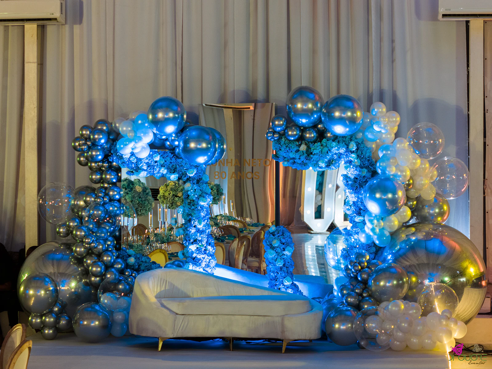 Palco de frente, com arcos cobertos de flores azuis e balões prateados e azuis, o chaise longue branco e, ao fundo, um espelho com letras douradas e o 80 luminoso