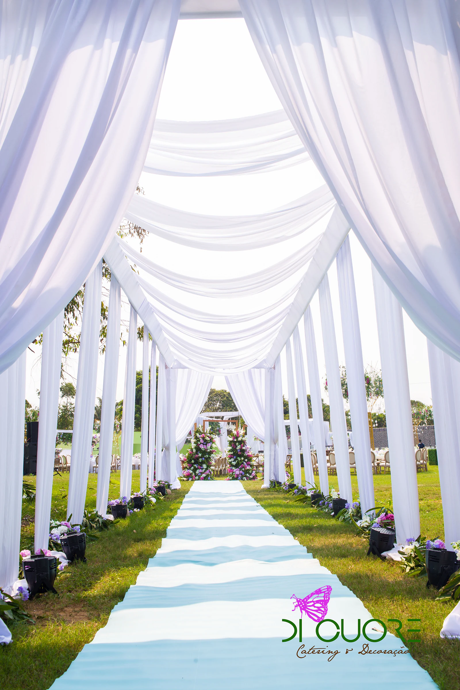 Avenue of white drapes and fabric pillars over a pale blue carpet, with two towers of flowers at the far end, on the lawn