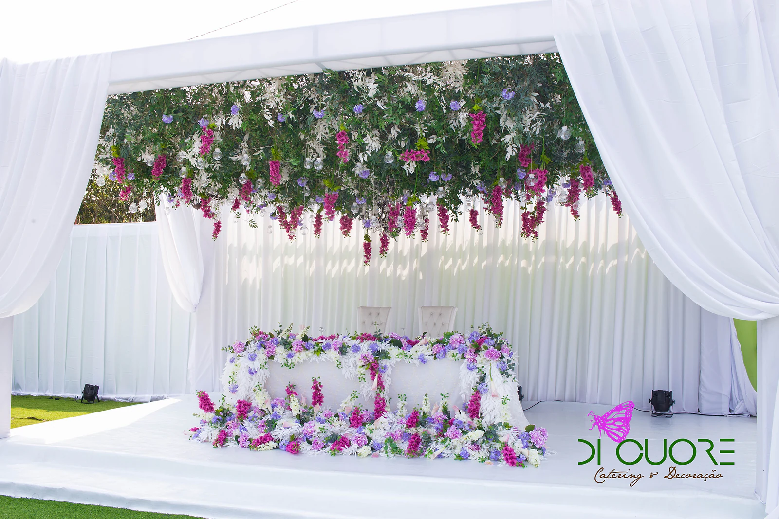 Mesa com duas cadeiras sob um pórtico de cortinados brancos, rodeada de flores fúcsia, lilases e brancas, com folhagem e cachos fúcsia suspensos