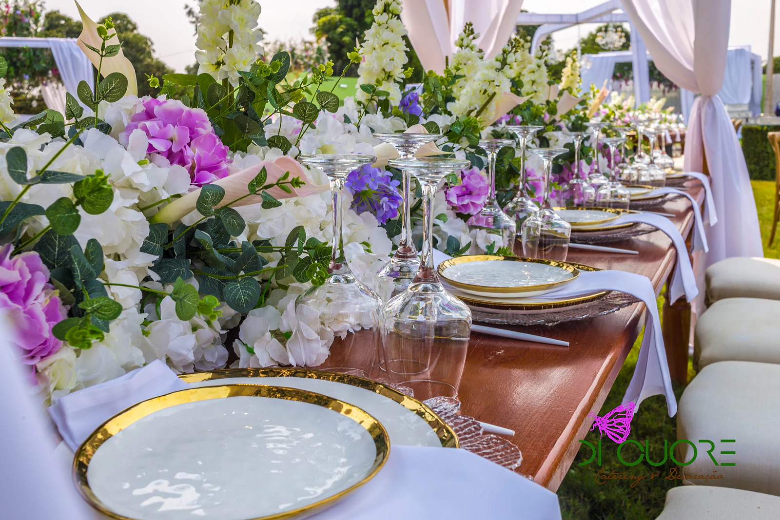 Prato branco de rebordo dourado sobre uma mesa corrida de madeira, com guardanapo branco, copos de pé alto e um ramo de hortênsias e flores brancas