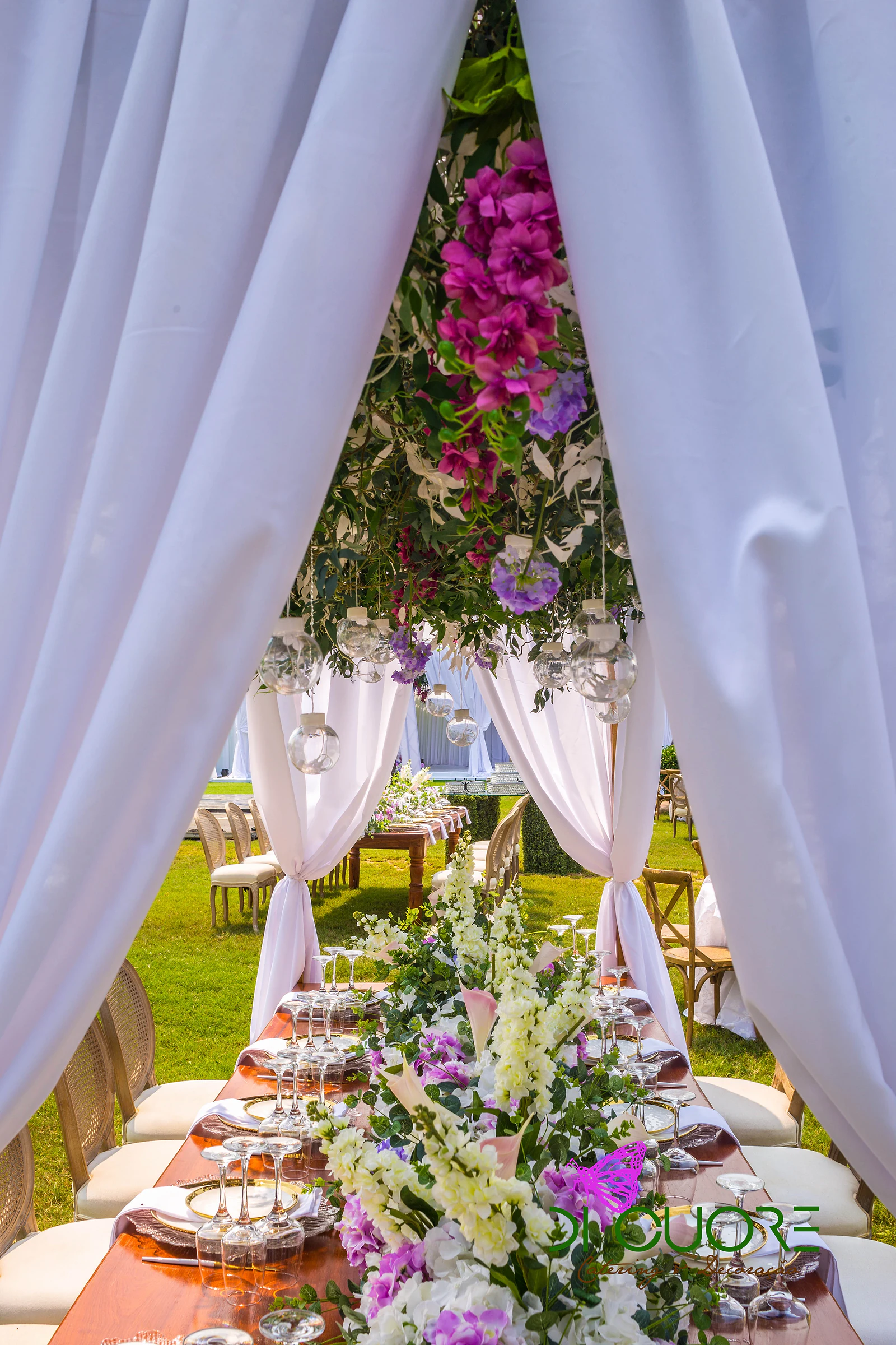 Cortinados brancos a emoldurar uma mesa corrida de madeira, com um caminho de flores brancas e lilases e uma cobertura de folhagem e flores fúcsia suspensas