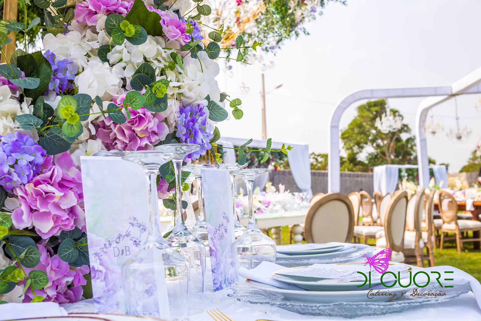 Menu em papel translúcido com as iniciais D&B, entre copos e pratos de vidro, junto a um ramo de hortênsias lilases e cor-de-rosa e folhagem