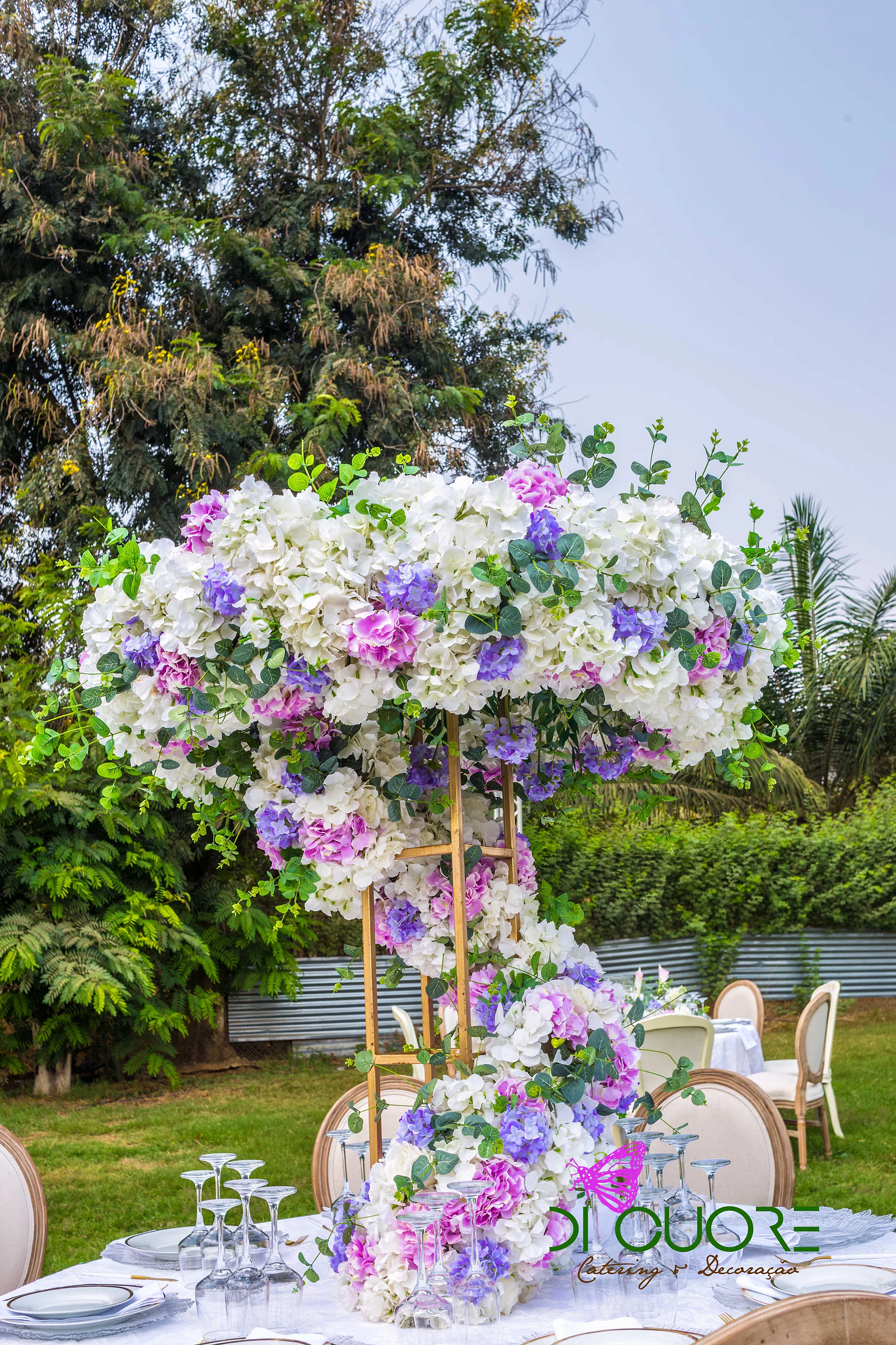 Centro de mesa alto em estrutura dourada, coberto de hortênsias brancas, lilases e cor-de-rosa e folhagem, com copos e pratos em primeiro plano
