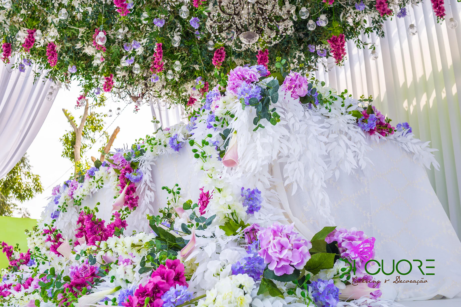 Pormenor da mesa sob a cobertura de flores: massa de hortênsias, flores fúcsia e ramos brancos sobre a toalha branca