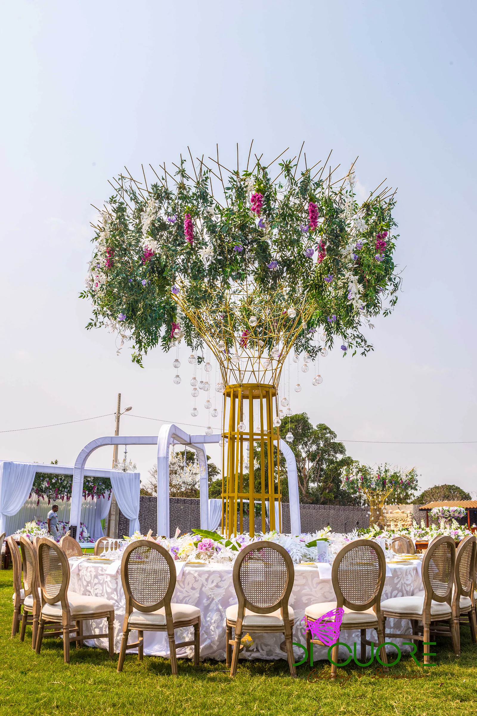 Árvore de metal dourado com uma copa de folhagem, flores fúcsia e bolas de vidro suspensas, sobre uma mesa redonda de toalha rendada e cadeiras de palhinha