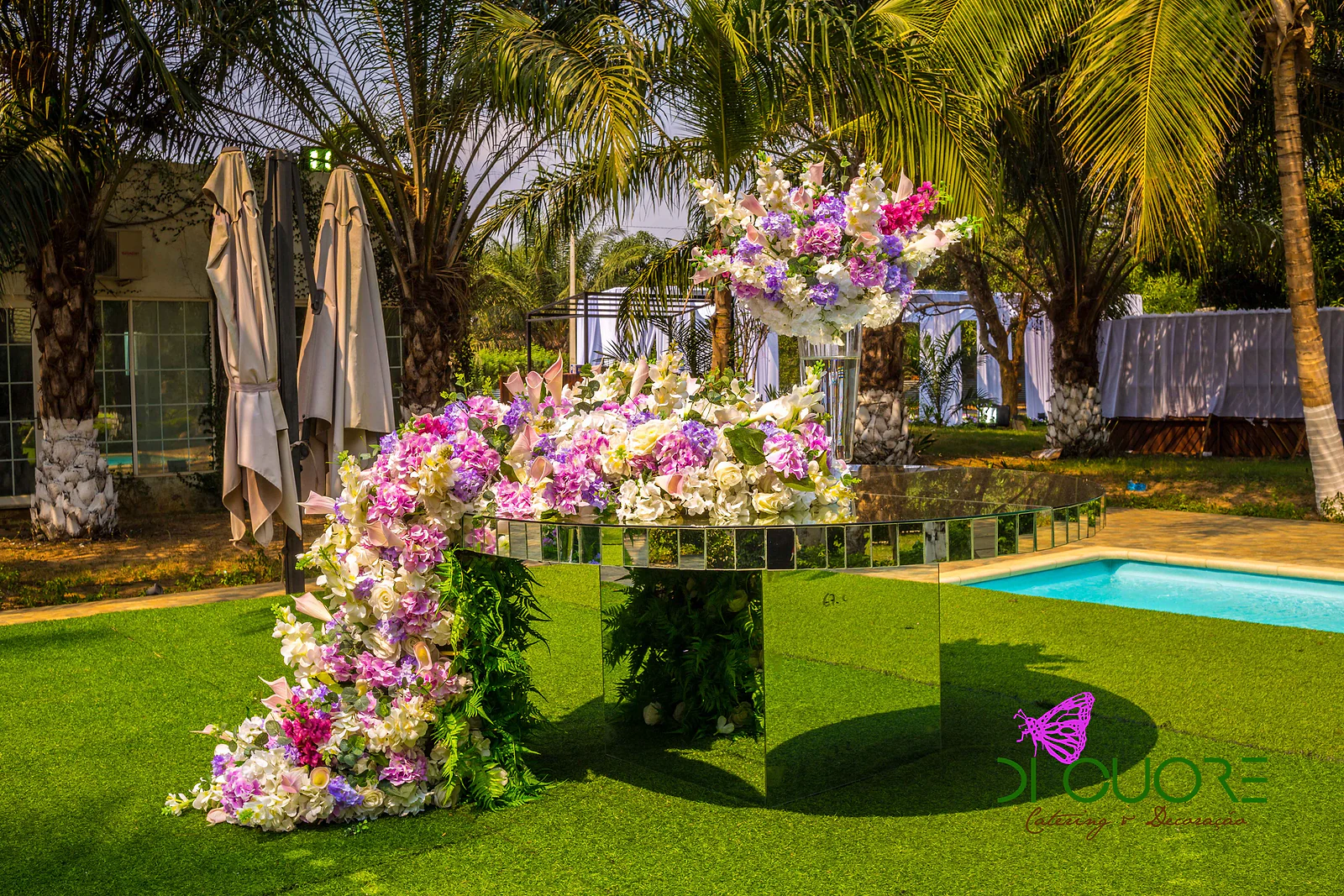 Pedestal de espelhos com uma cascata de flores lilases, cor-de-rosa e brancas que desce até à relva, junto à piscina e entre palmeiras