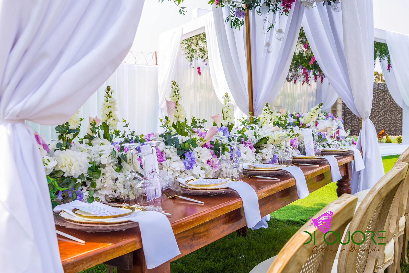 Mesa corrida de madeira entre cortinados brancos atados, com flores brancas e lilases, pratos de rebordo dourado e guardanapos brancos
