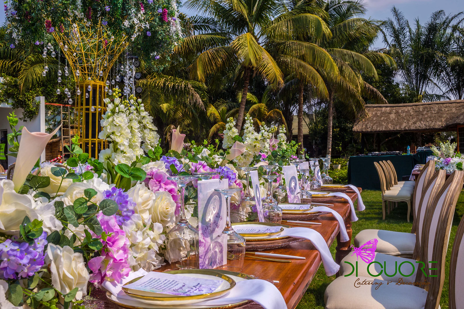 Mesa corrida de madeira com caminho de flores brancas e lilases, pratos de rebordo dourado e menus, e uma árvore dourada ao fundo