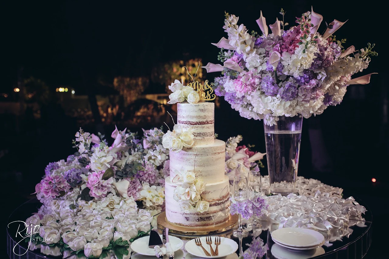 Bolo de três andares com rosas brancas, sobre um espelho coberto de flores lilases e brancas, com um grande ramo num vaso alto, à noite
