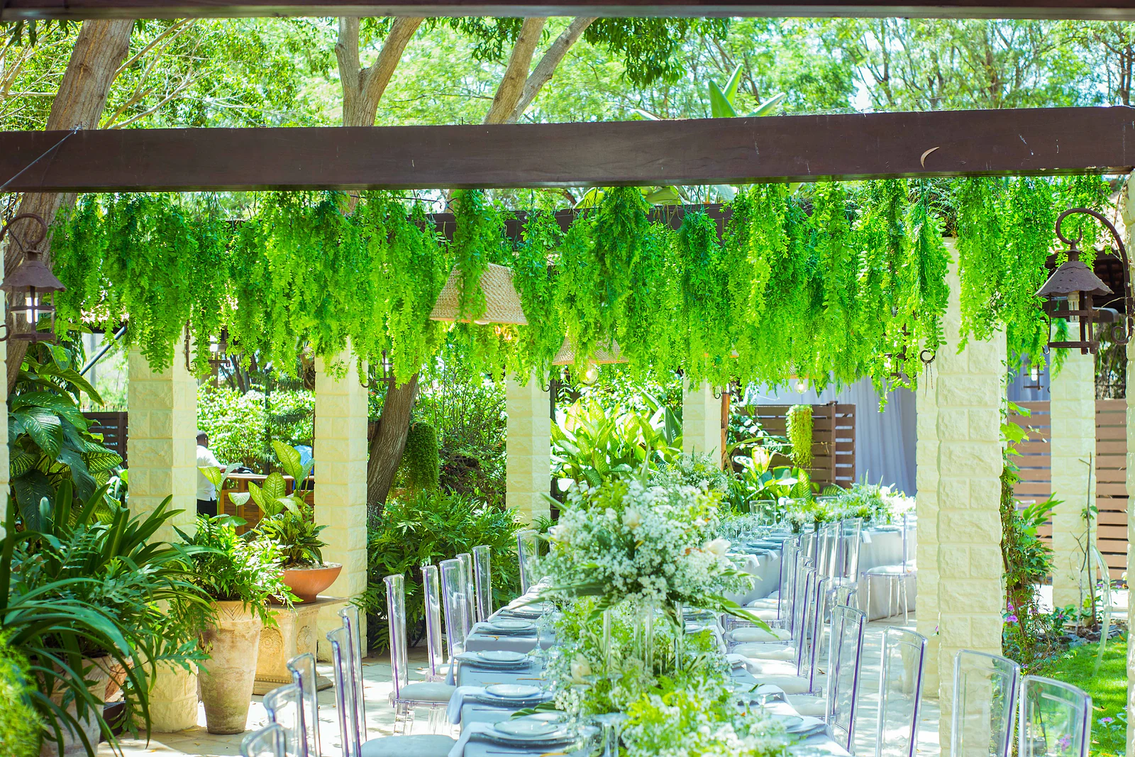 Stone pergola with hanging foliage above a long laid table, white flowers along the centre and clear chairs