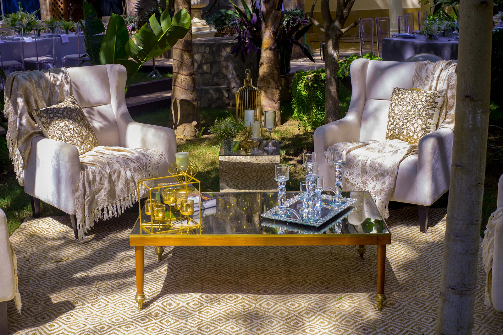 A corner with two pale armchairs, fringed throws and embroidered cushions around a mirrored coffee table with a gold frame