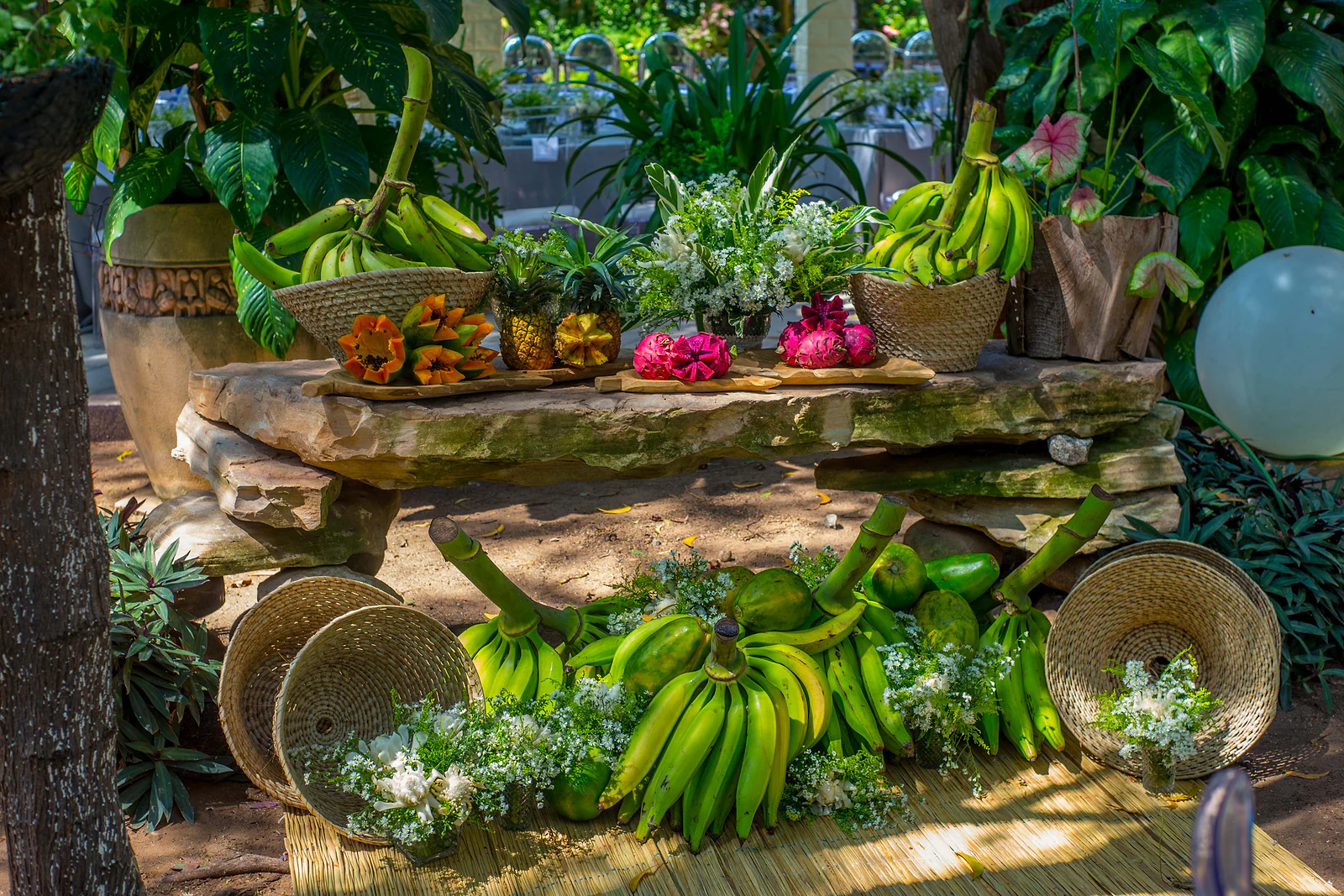 Stone ledge with baskets of bananas, pineapples and dragon fruit among white flowers, on a straw mat