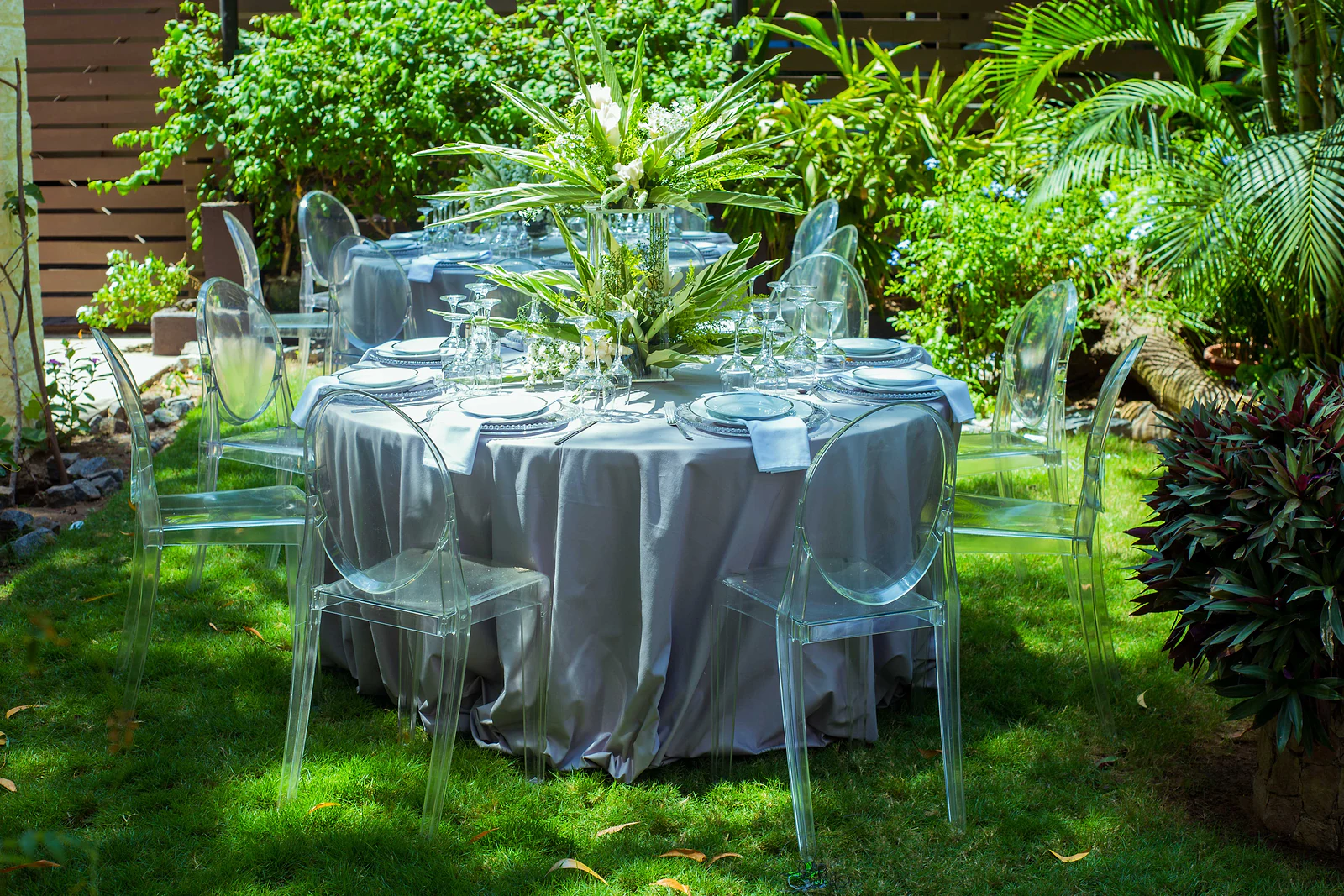 Round table with a grey cloth and a tall arrangement of foliage and white flowers, on the lawn among palms and plants, surrounded by clear chairs