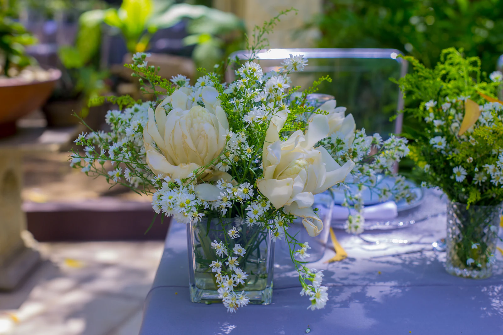 Square glass vase of white roses and tiny gypsophila flowers on a lilac tablecloth in garden light