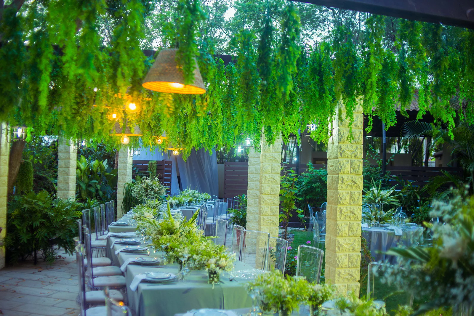 Long table at dusk beneath hanging foliage and lit straw lampshades, between pillars of yellow stone