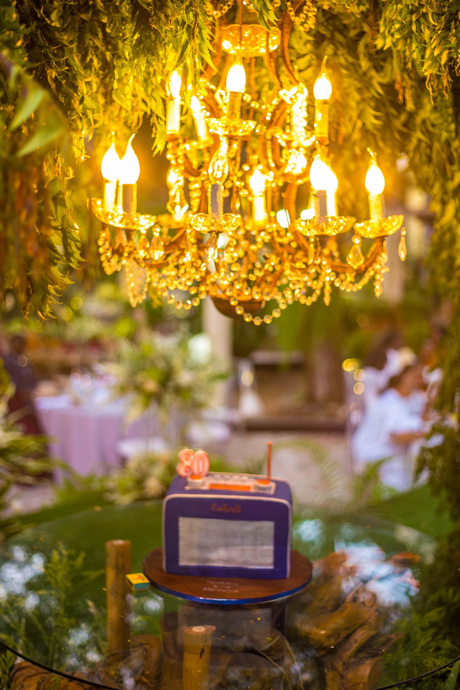 A lit golden chandelier among hanging foliage and, below it, the blue radio-shaped cake with the 80 candle on a glass table