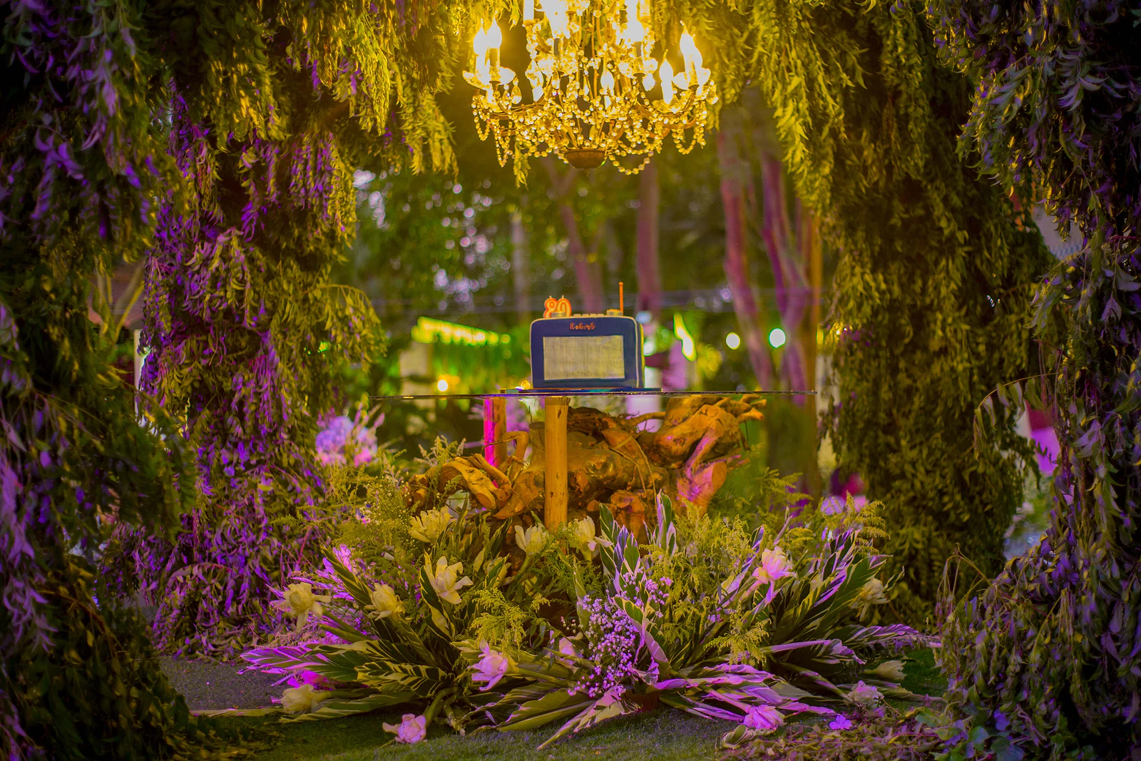 Cenário ao fim da noite sob folhagem pendente e luz lilás, com flores cor-de-rosa ao chão e, sobre uma mesa de vidro, o bolo em forma de rádio sob um lustre dourado