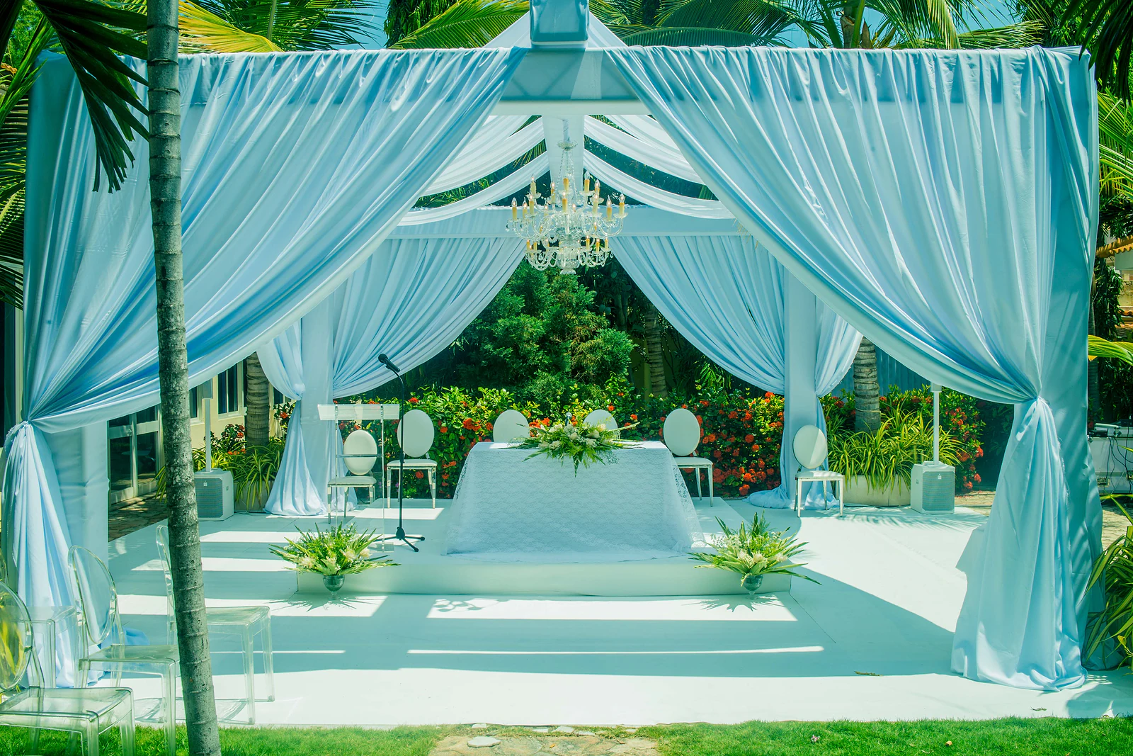 Pavilion of pale blue drapes with a crystal chandelier, a table dressed in white at the centre, planters of foliage and clear chairs