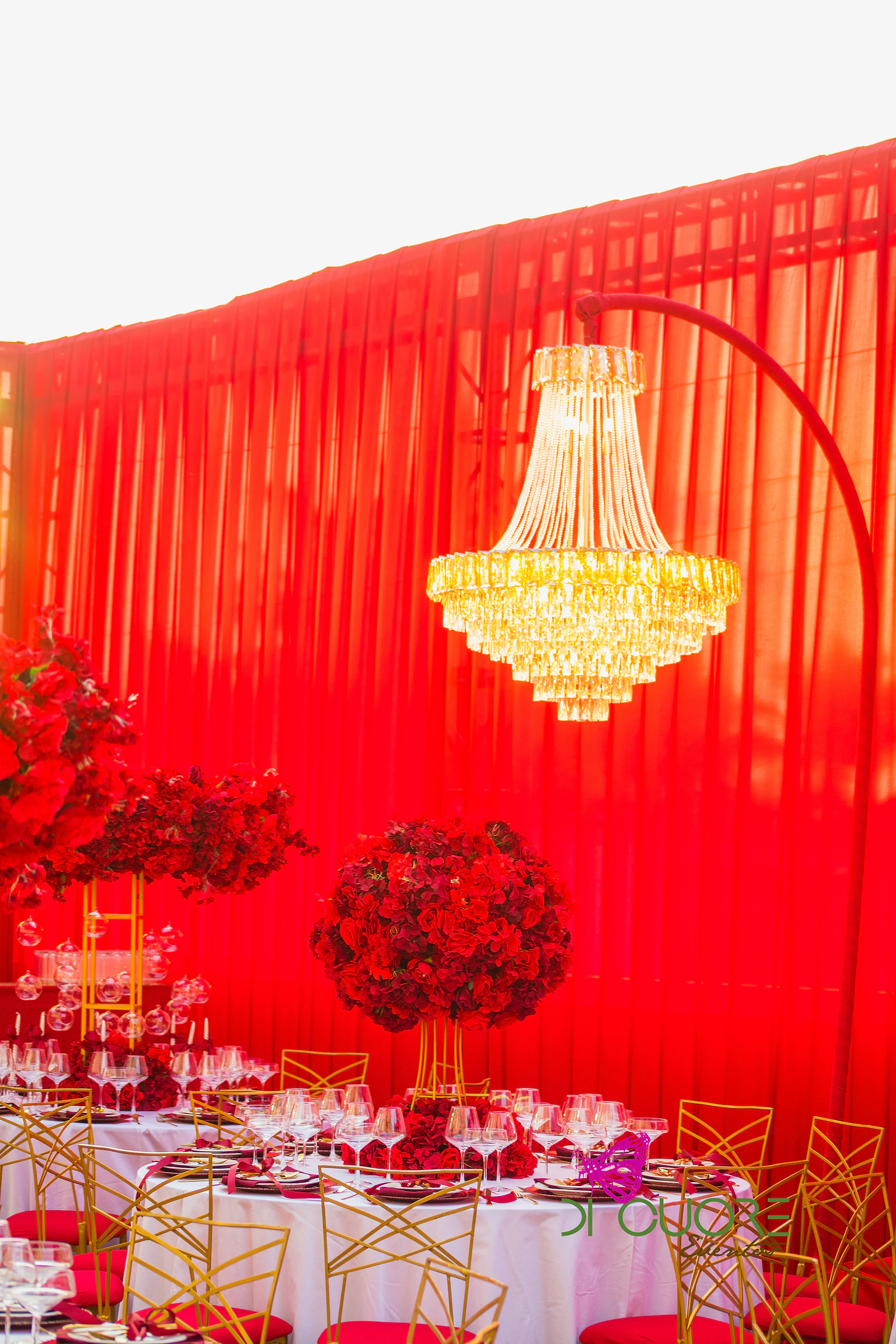 Lustre de cristal dourado diante de cortinados vermelhos, com esferas de rosas vermelhas sobre as mesas e cadeiras douradas