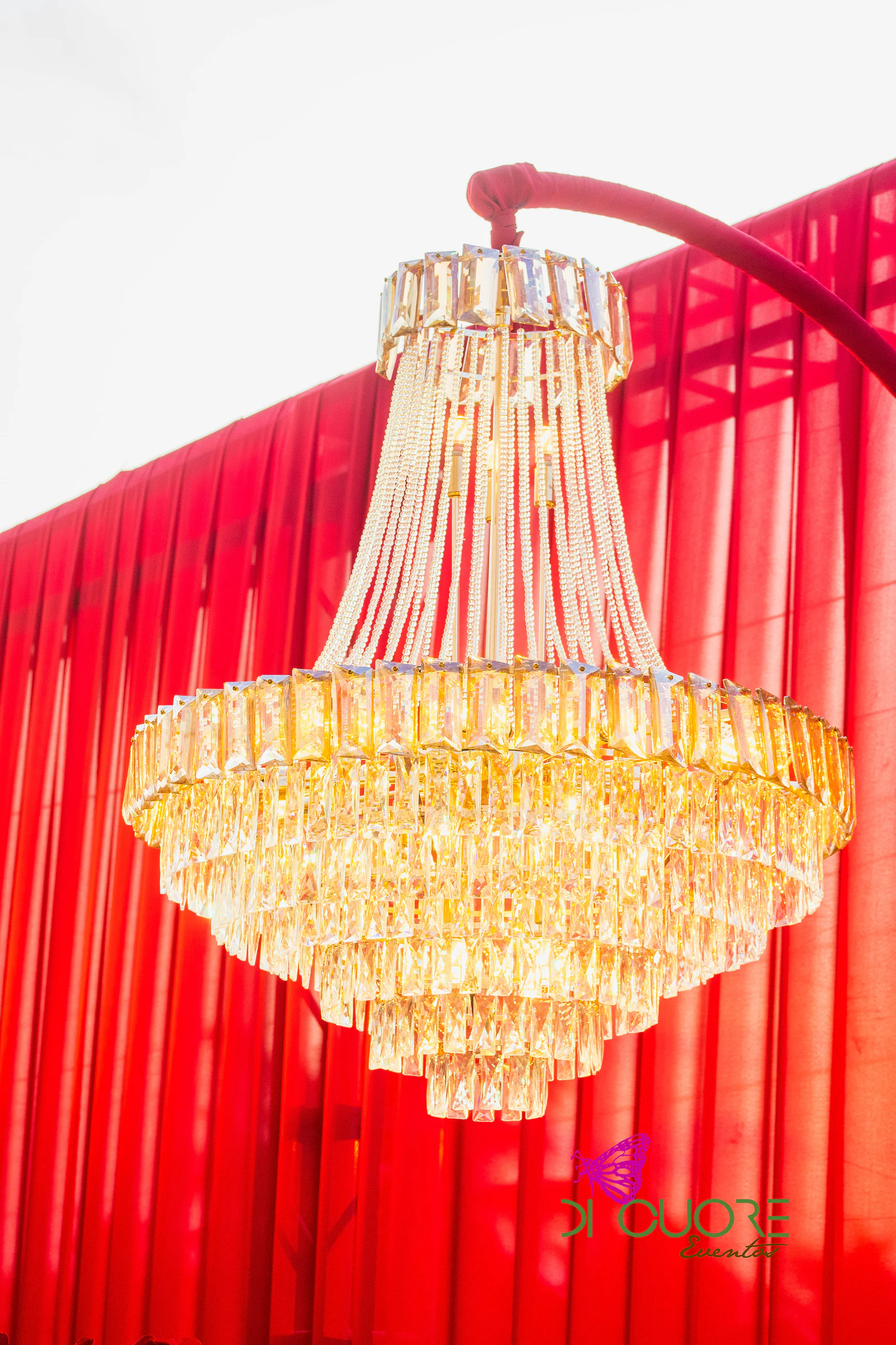 Pormenor de um lustre de cristal dourado pendurado de um arco vermelho, sobre cortinados rubros