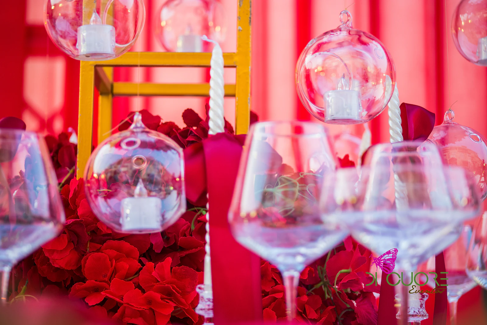 Esferas de vidro com velas suspensas sobre rosas vermelhas, copos de cristal e fitas carmim