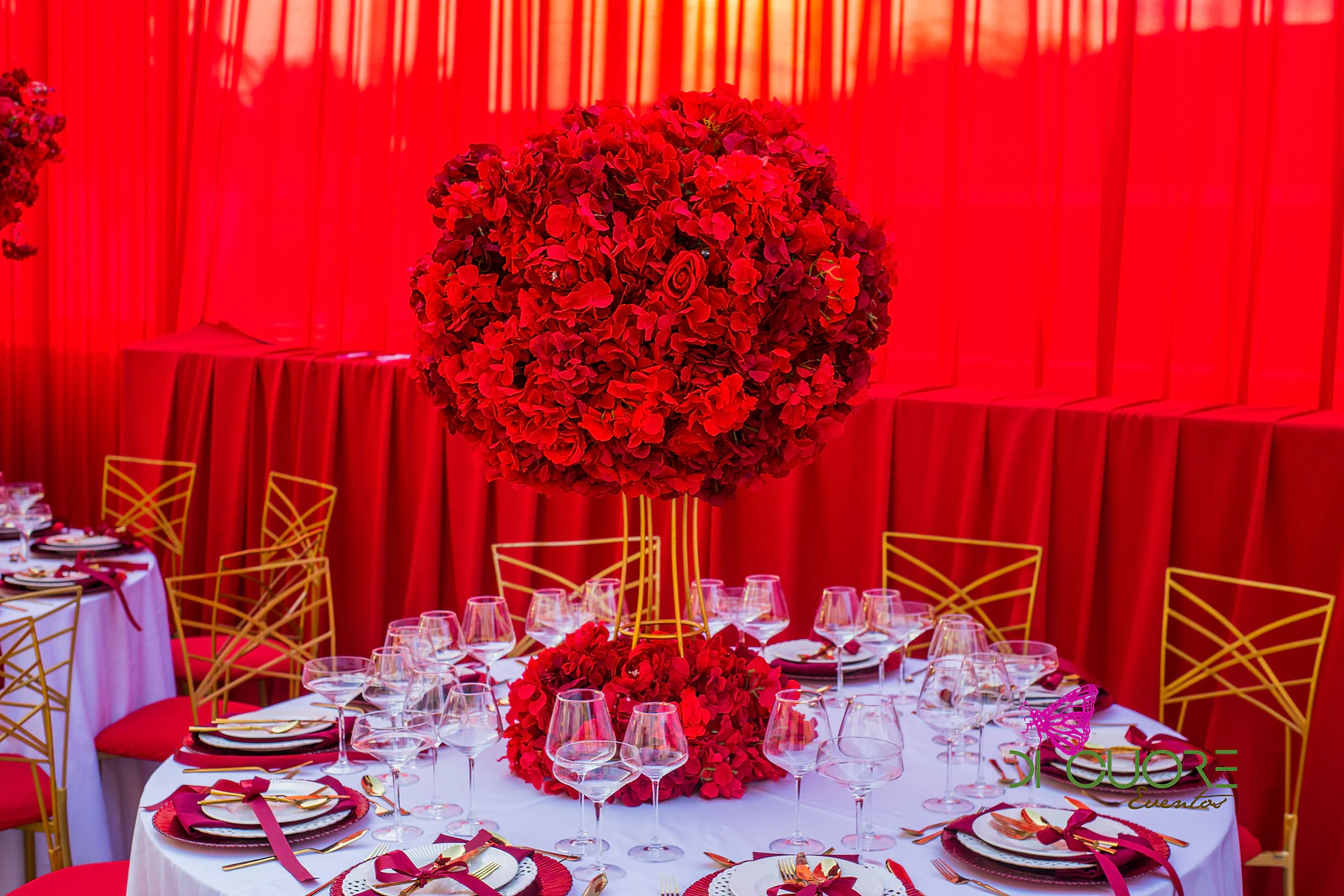 Mesa redonda com esfera de rosas vermelhas sobre pedestal dourado, copos de cristal e cadeiras douradas diante de um cortinado vermelho