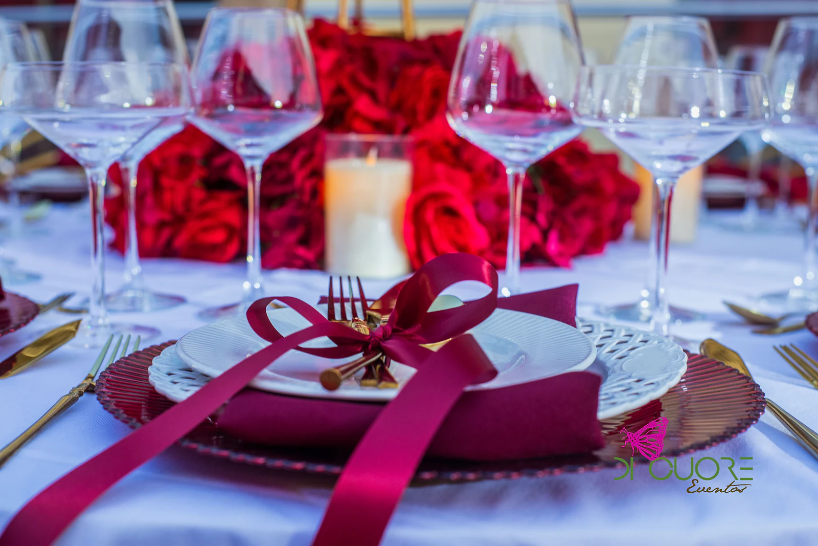 Lugar à mesa com laço de fita carmim, copos de cristal e uma vela entre rosas vermelhas
