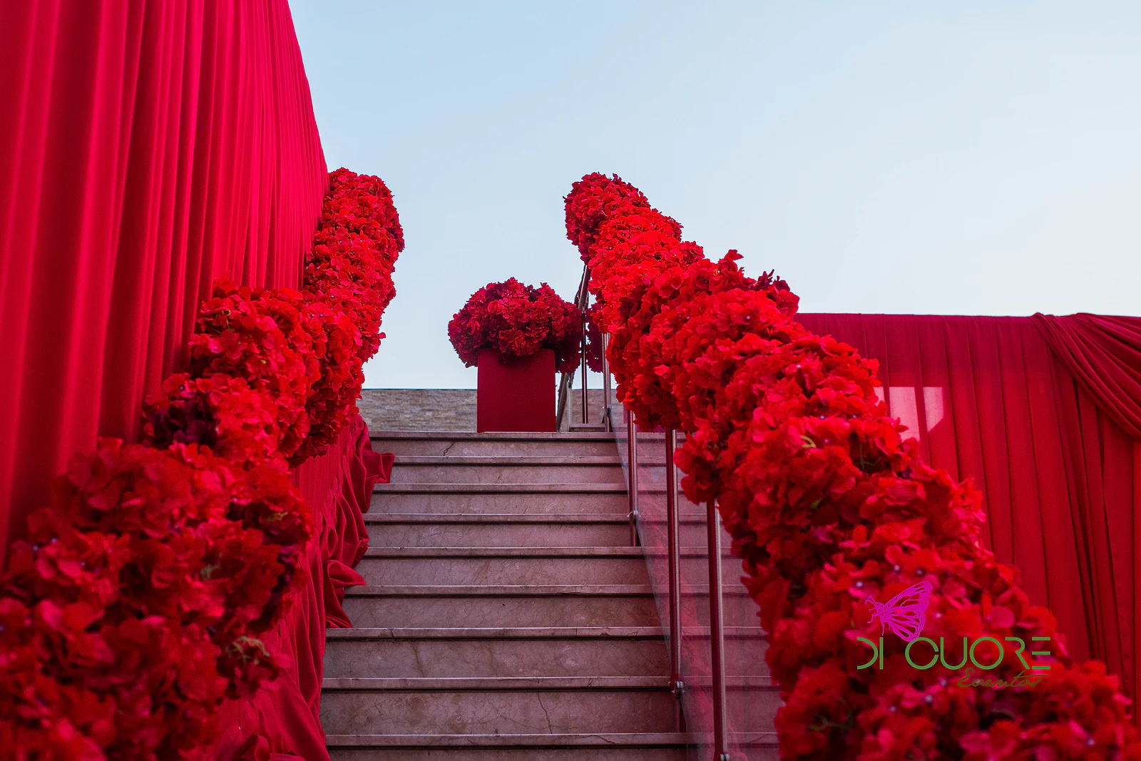 Escadaria ladeada por guirlandas de rosas vermelhas e cortinados rubros, sob o céu ao fim do dia