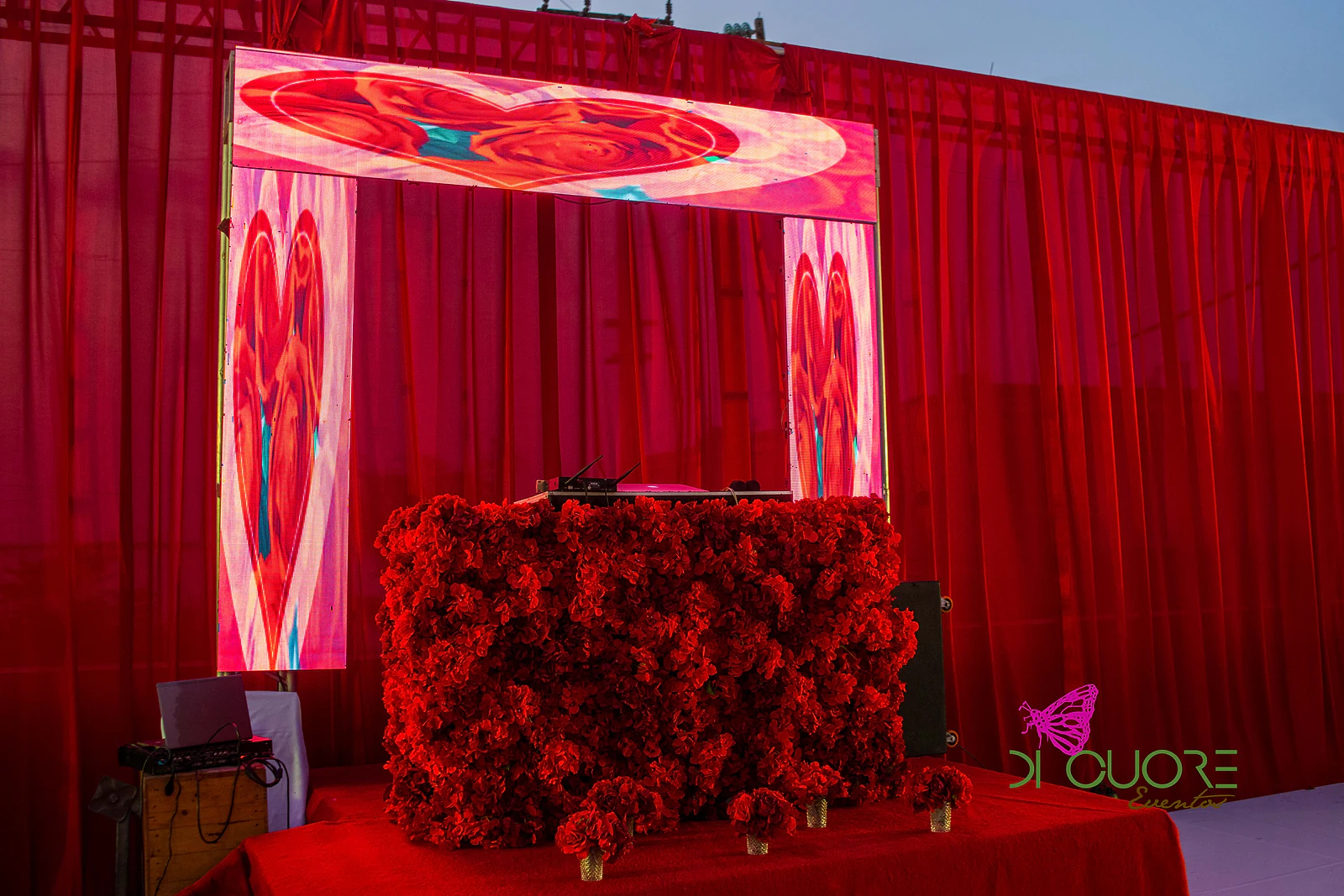 Mesa de DJ coberta de rosas vermelhas diante de ecrãs de LED com desenhos de rosas e corações, entre cortinados vermelhos