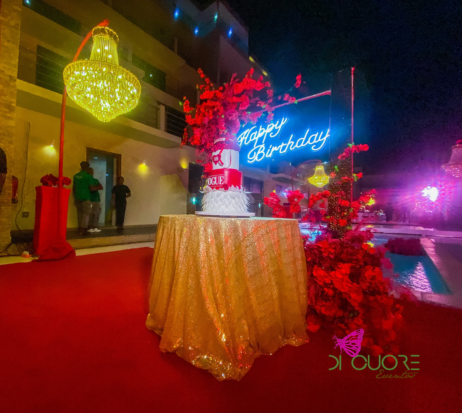 Bolo de dois andares sob um letreiro de néon azul com Happy Birthday, numa mesa de toalha dourada com cascata de rosas vermelhas e tapete vermelho