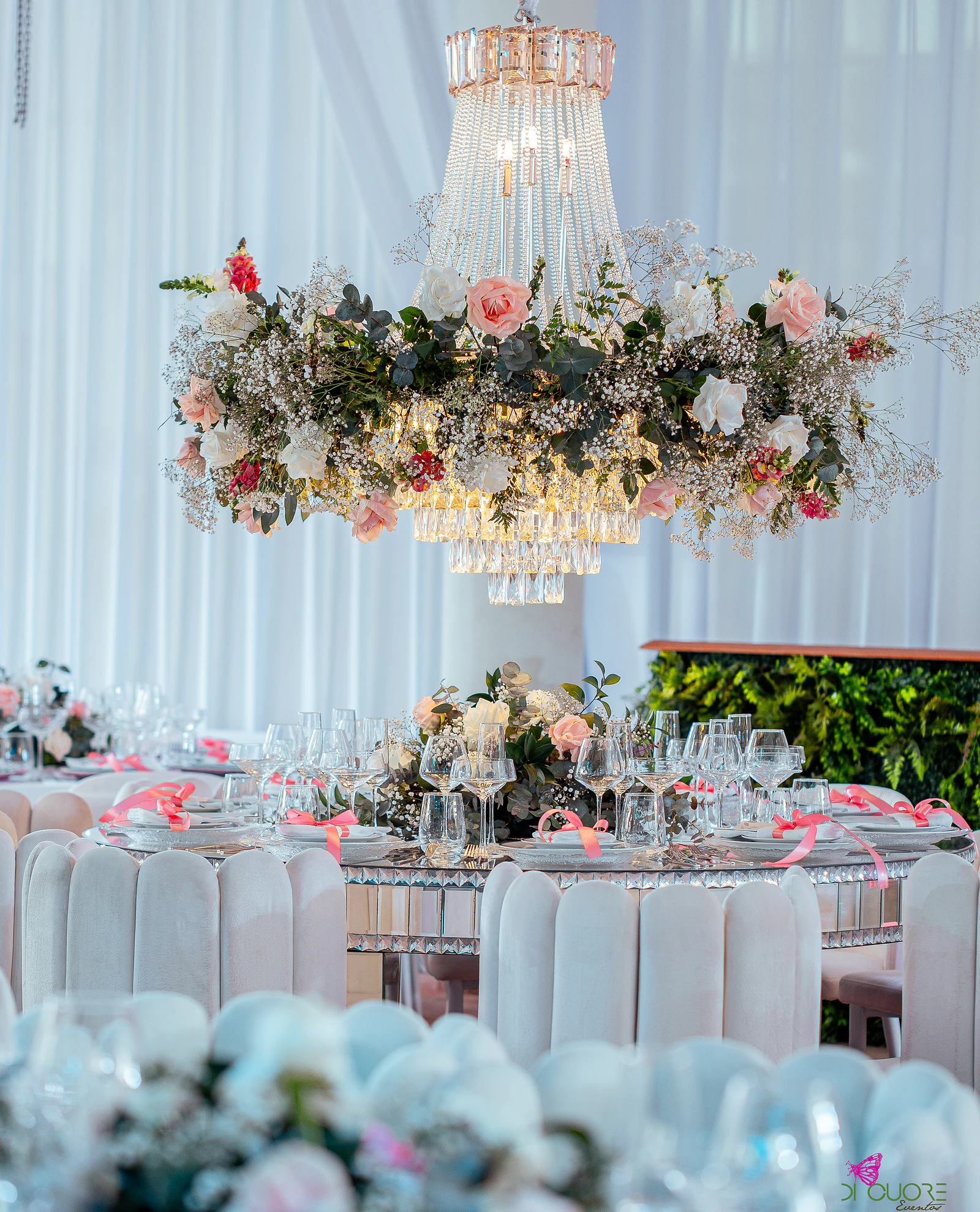 Lustre de cristal com uma coroa de rosas, gisófila e folhagem pendurado sobre uma mesa redonda com fitas cor-de-rosa, entre cortinados brancos