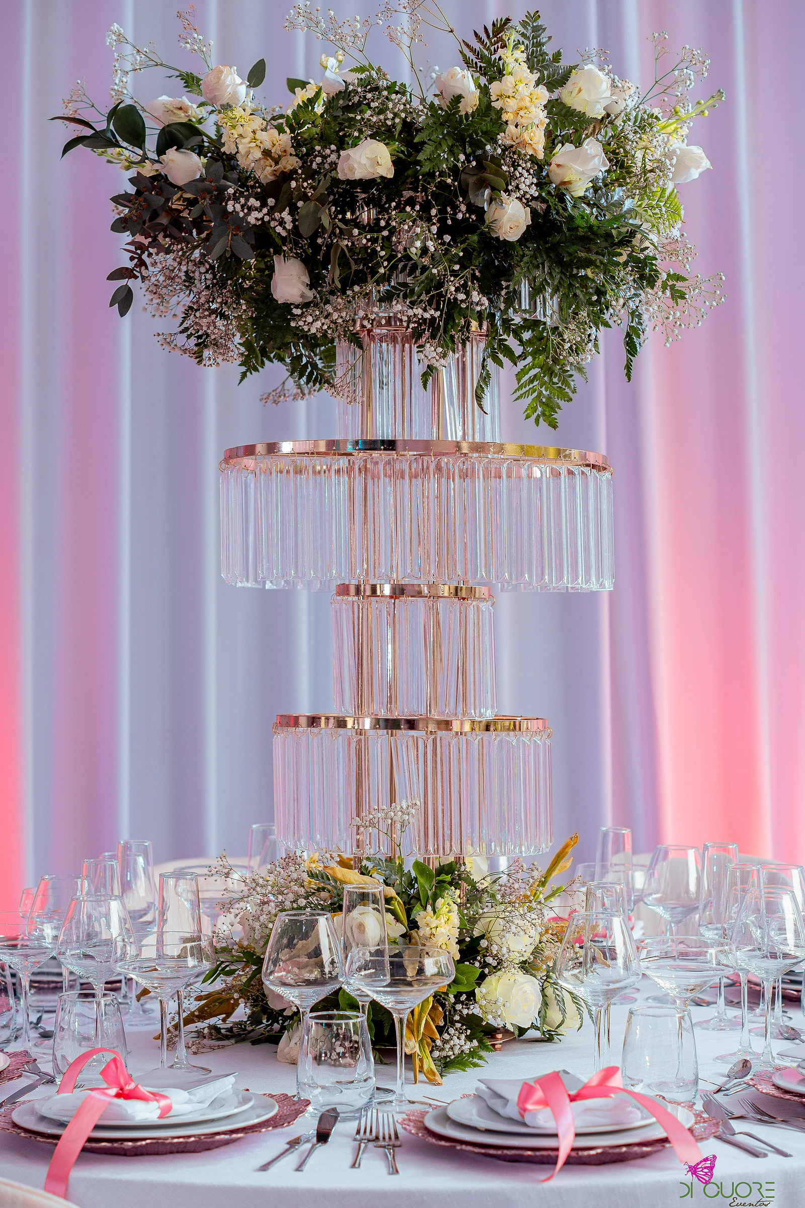 Coluna de cristal em três andares com bordos dourados e uma coroa de rosas brancas, folhagem e gisófila, sobre uma mesa posta com fitas cor-de-rosa