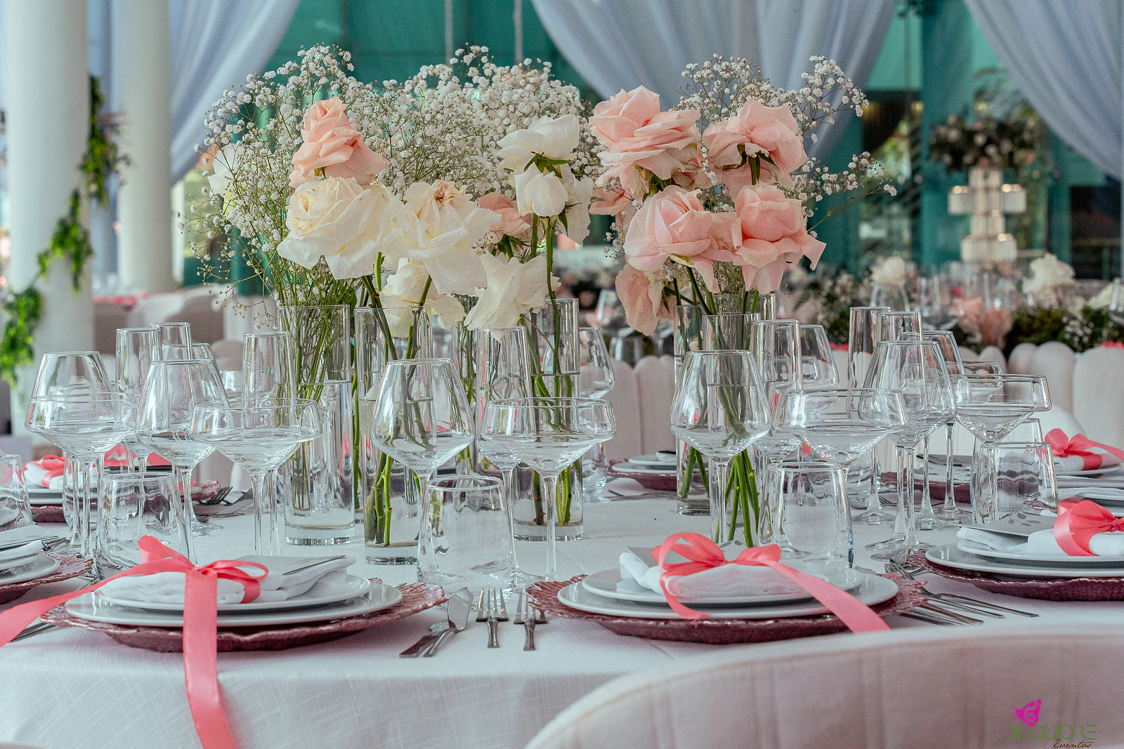 Jarras altas com rosas cor-de-rosa e brancas e gisófila sobre uma mesa posta, com fitas cor de coral nos guardanapos