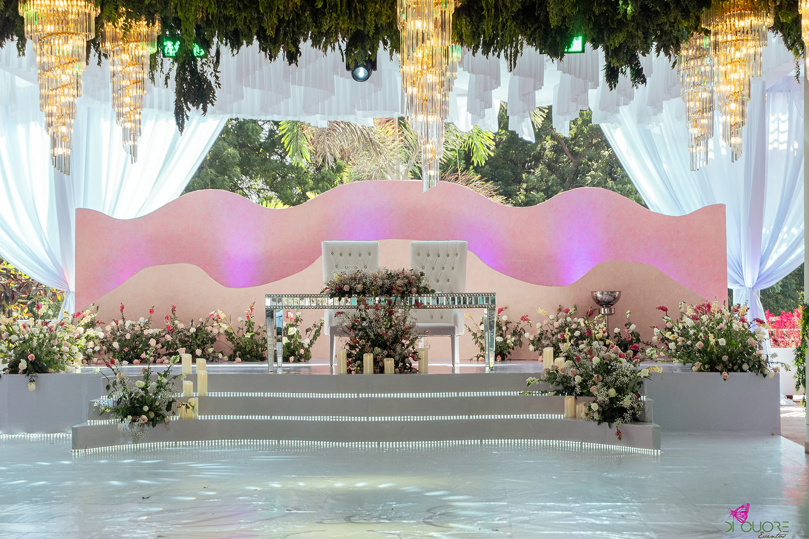 Palco de linhas onduladas em tons de rosa com dois tronos brancos e uma mesa de espelho, rodeado de flores, sobre degraus iluminados