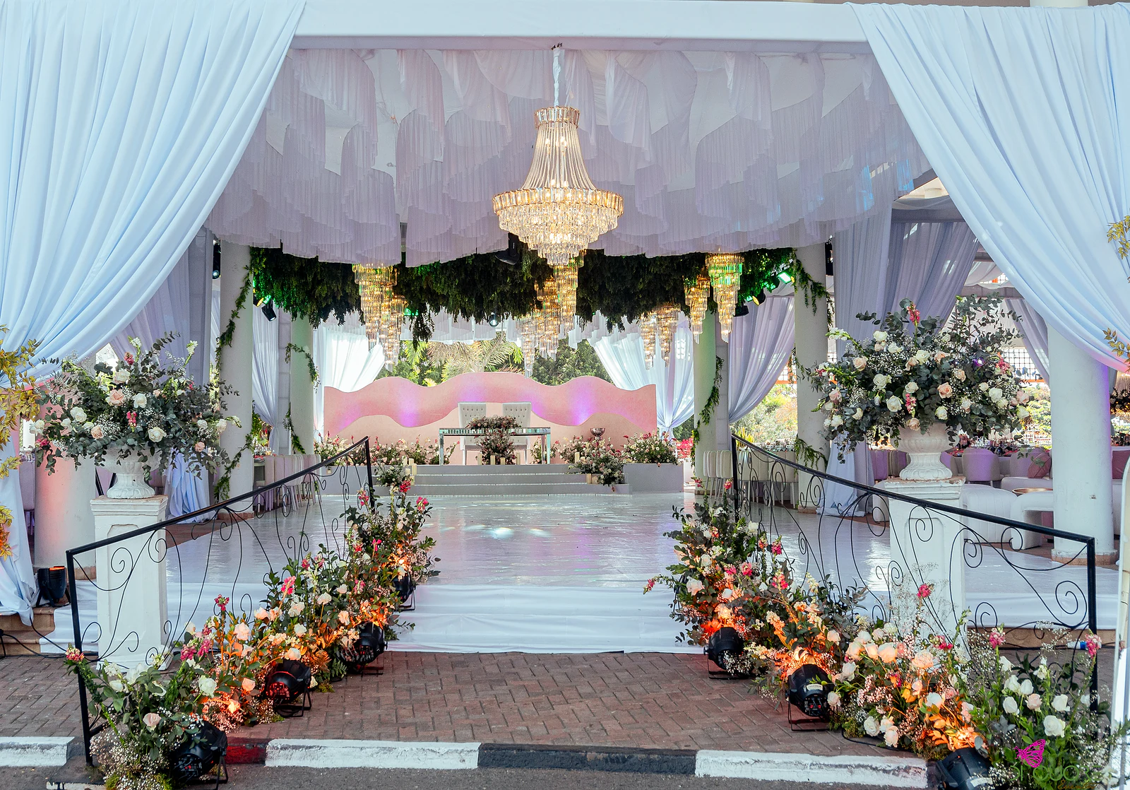 Entrada do salão com cortinados brancos, lustre de cristal, folhagem suspensa e ramos de flores de cada lado do caminho, em direcção ao palco cor-de-rosa